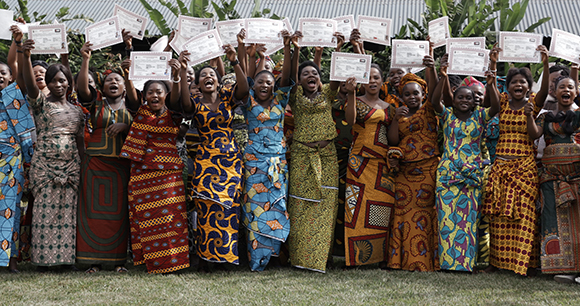 A large group of women in vibrant, patterned African attire smiling and holding up white certificates.