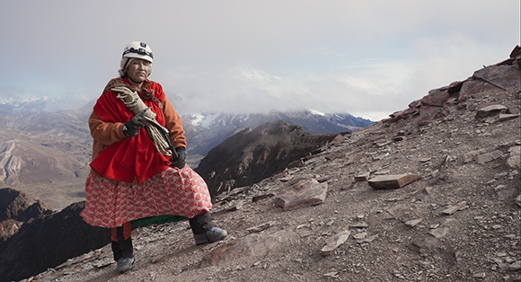 A person wearing a red tunic, patterned skirt, and white climbing helmet stands on a steep, rocky mountainside.
