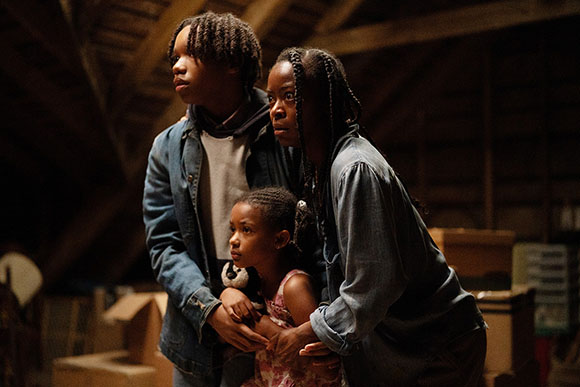 A young man, woman, and small girl look ahead with worried expressions in a dark, wooden attic.