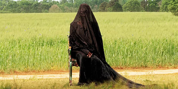 A person draped in a long, black lace veil sits on a chair in a grassy field.