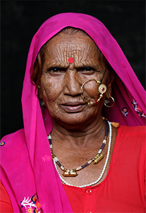 Close-up portrait of an elderly woman wearing a bright pink headscarf, a large gold nose ring, and beaded necklaces.