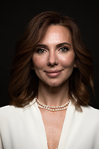 A woman with wavy brown hair, wearing a white blazer and a double-strand pearl necklace against a black background.