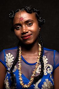 A young woman with dark braided hair, a red sun symbol on her forehead, and a shell necklace.