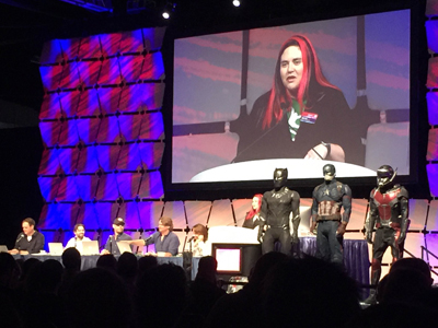 A panel of people sit at tables on a stage, with Black Panther, Captain America, and Ant-Man costumes standing in front of th