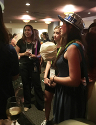 Several women are gathered in a room, with some wearing hats and lanyards. One woman in the foreground wears a blue dress and