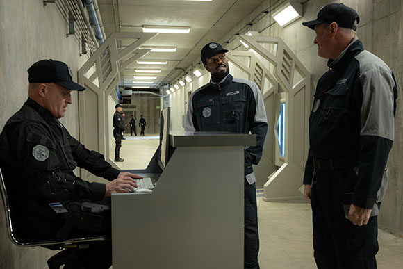 Three men in dark tactical uniforms stand in a brightly lit, modern concrete corridor.