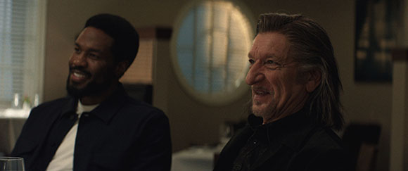 A Black man and an older man with long hair sit side-by-side, both smiling.