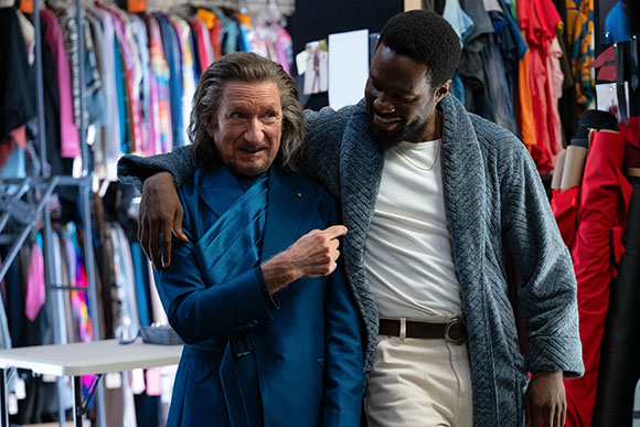 A man in a blue suit and a man in a grey robe embrace in a room filled with colorful clothing.
