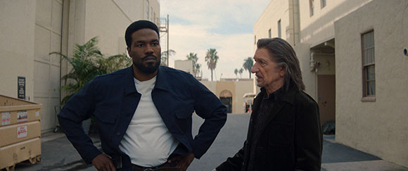 A man in a blue jacket and a man in a black coat stand in an outdoor alleyway.