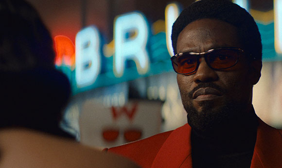 A man with a beard and tinted glasses wearing a red jacket looks toward a person in the foreground.