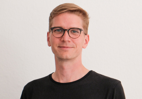 A man with short blond hair and black-rimmed glasses wearing a black shirt against a white background.
