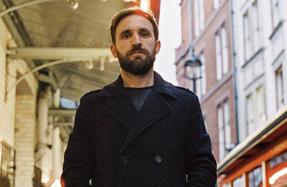 A bearded man wearing a dark double-breasted coat stands on a city street with buildings in the background.