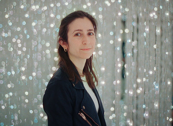 A woman with dark hair and gold hoop earrings looks toward the camera against a backdrop of blurred lights.