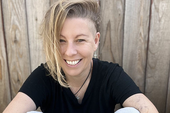 Smiling woman with blonde hair and a black t-shirt sitting before a wooden fence.