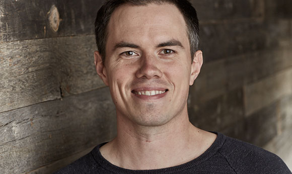 A man with short dark hair smiling against a rustic wooden plank background.