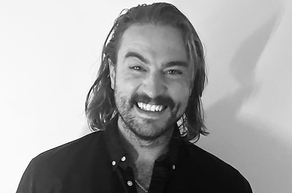 Black and white portrait of a smiling man with long hair and a beard wearing a dark shirt.