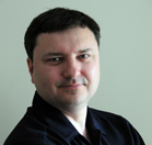 A man with short dark hair wearing a dark navy button-down shirt against a plain light background.