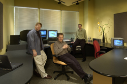 Three men posing in a modern office space with computer monitors and curved desks.