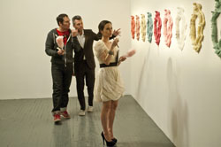 Three people stand in a white gallery looking at colorful, textured sculptures mounted on a wall.