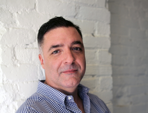 A man with short dark hair wearing a blue and white checkered shirt against a white brick wall.