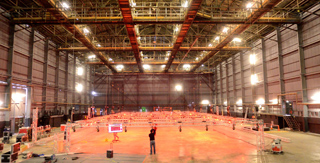 Interior view of a vast, empty industrial warehouse with high ceilings and bright lights.