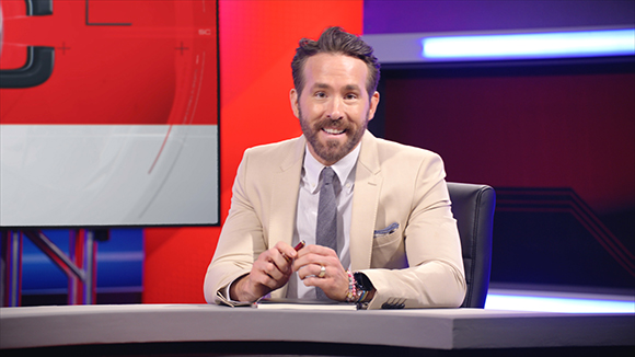 A man with a beard wearing a beige suit and tie sits at a desk in a studio.