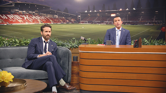 Two men in suits sit in a studio with a large, lit soccer stadium in the background.