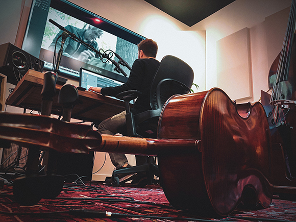 A man sits at a desk with a large monitor in a studio, viewed behind a large wooden cello.