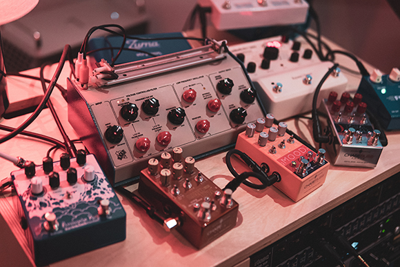 Multiple colorful electronic music synthesizers and effect pedals with knobs and cables on a wooden surface.