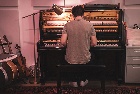 A person seen from behind playing a black upright piano under a warm lamp.