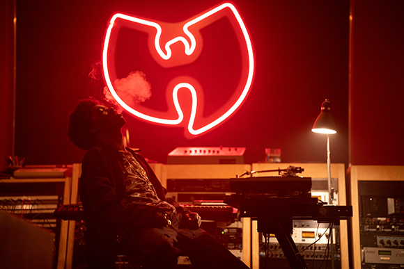 A person exhales smoke in a dark studio lit by a large red neon Wu-Tang Clan logo and a desk lamp.