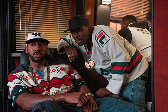 Two men wearing baseball caps and heavy gold chains sit inside a vehicle with window blinds.