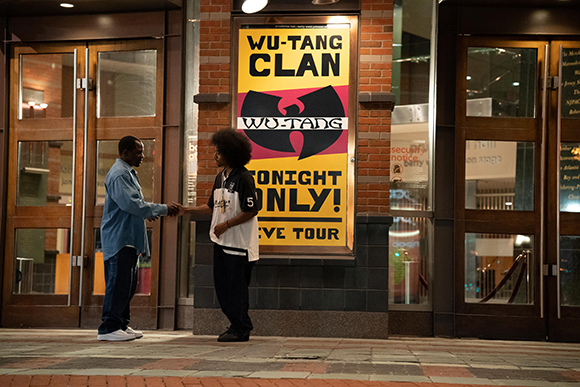 Two men shake hands on a sidewalk in front of a large Wu-Tang Clan concert poster.