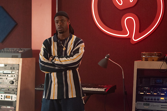 A man in a striped polo shirt and black cap stands with arms crossed in a music studio.