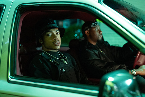 A man in a black bucket hat and silver chain looks out a car window while another man sits in the driver's seat.