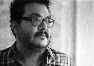 Black and white close-up portrait of a man with dark curly hair, glasses, and a beard wearing a plaid shirt.