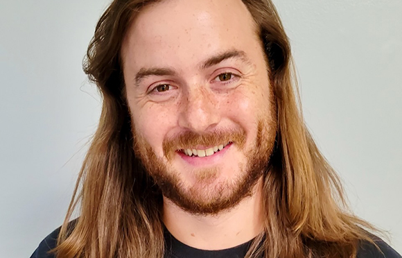 A smiling man with long brown hair, freckles, and a beard against a light blue background.