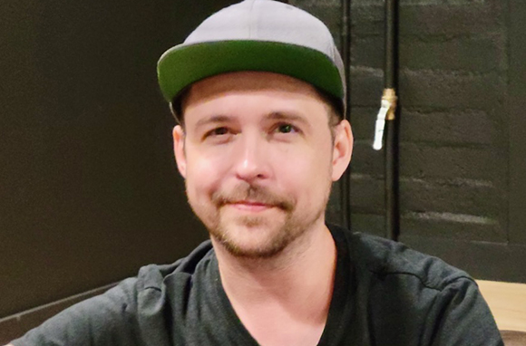 A man with a light beard and a green and grey baseball cap smiling slightly.