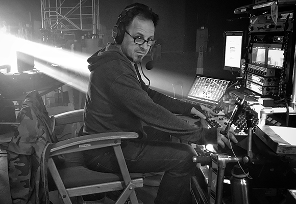 A man in a hoodie and glasses sits at a desk with monitors and audio equipment in a dark studio.