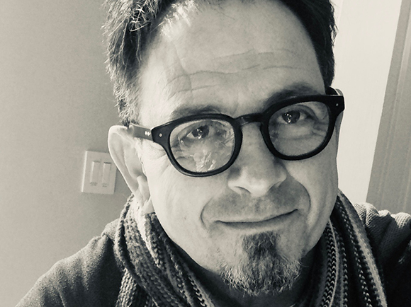 Black and white close-up portrait of a man with glasses, a goatee, and a textured scarf.