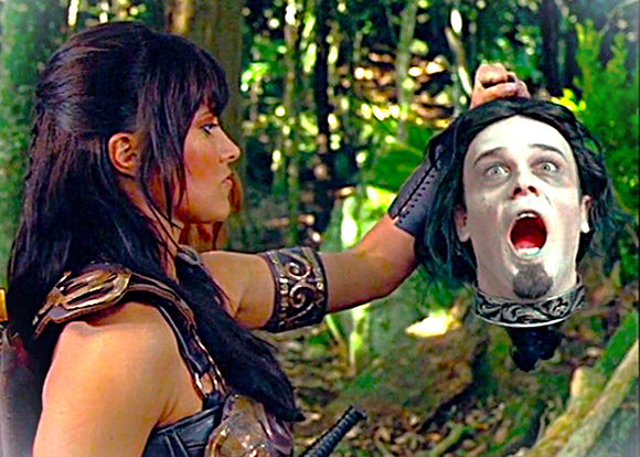 A woman with dark hair holds a severed head by its hair in a forest setting.
