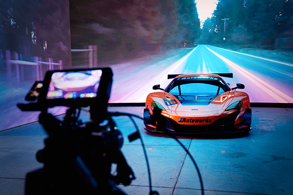 A professional camera captures an orange race car positioned before a large screen displaying a digital road.