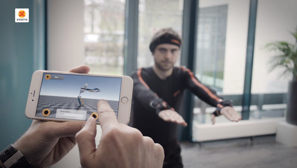 Hands hold a smartphone displaying a digital avatar while a man in a black motion capture suit stands in the background.