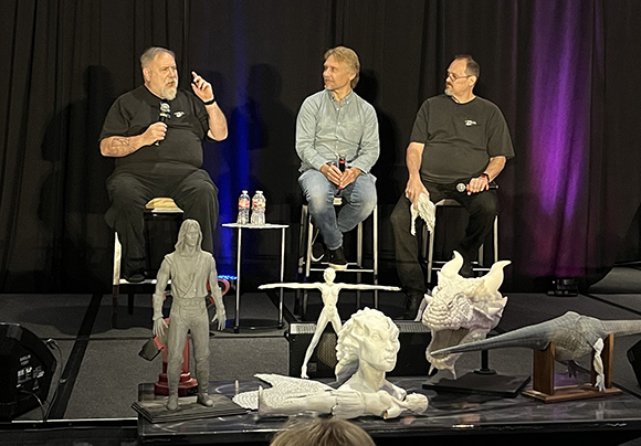 Three men sit on stools on a stage behind several white and grey sci-fi character sculptures.