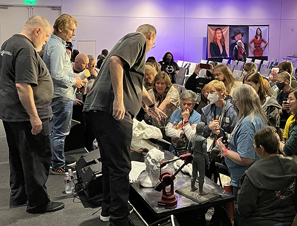 A group of people stand around a table with a white bust and a small dark figurine at an indoor event.