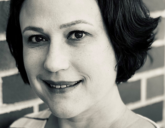 Black and white close-up portrait of a smiling woman with short dark hair against a brick wall.