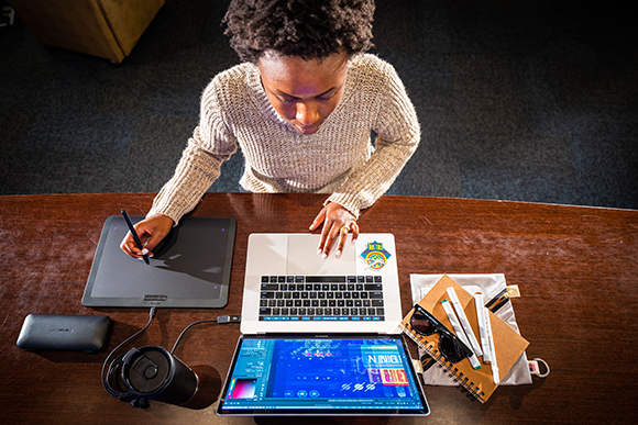 A person uses a stylus on a drawing tablet next to an open laptop and a notebook on a wooden table.