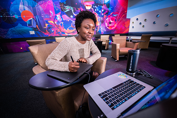 A person sits at a table using a stylus on a drawing tablet next to an open laptop.