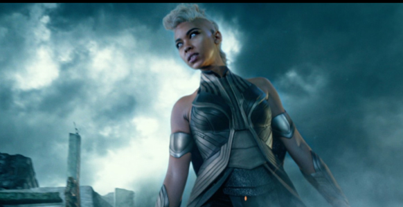 A woman with short blonde hair wearing silver armor stands against a dark, cloudy sky.