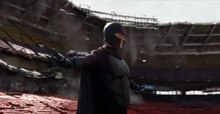 A person in a dark, armored superhero suit with a cape stands amidst the rubble of a destroyed stadium.